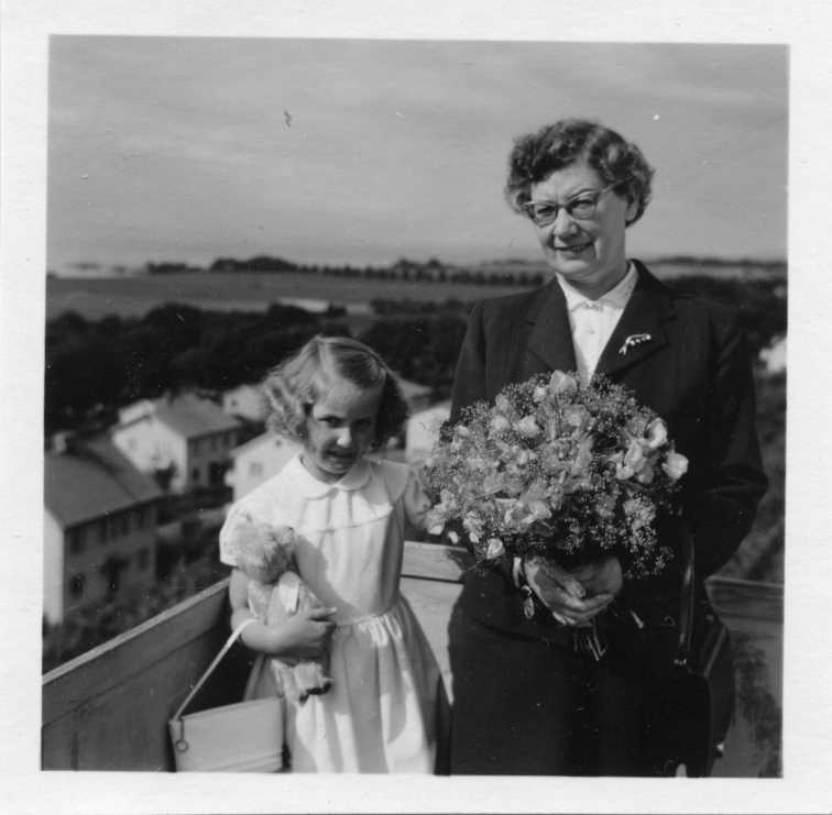 Sonja Fong och flickan Gunilla Fong står på en balkong i Gränna. Gunilla har en ljus klänning på sig, en handväska på armen och en nalle i famnen. Sonja håller en bukett blommor i händerna.