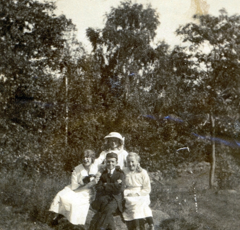 Tre flickor (troligen sommargäster) och Gunnar Karlsson, Vommedal  Östergård "Skräddarns" (1901 - 1979) står på en gräsmatta, Vommedal 1920-tal. Relaterat motiv: A0013.