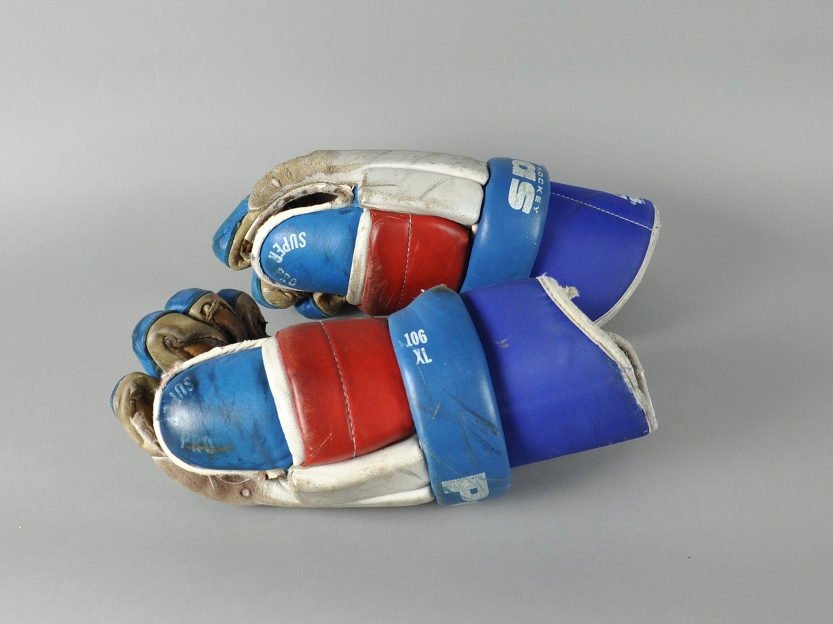 Et par Pallas ishockeyhansker av skinn i blått, rødt og hvitt. Hanskene er velbrukte og har flere hull på innsiden av hånden.