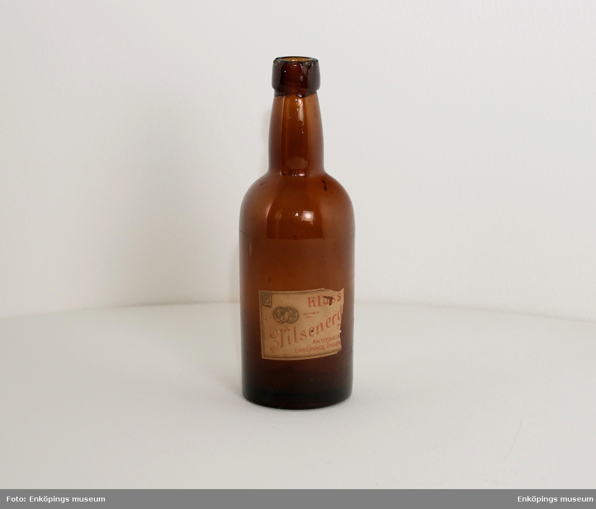 Brun flaska med etikett " Klass..., pilsnerd..., Aktiebola..., Enköpings Ångbr.... Märkt ibotten med " ÅRNAC"?.