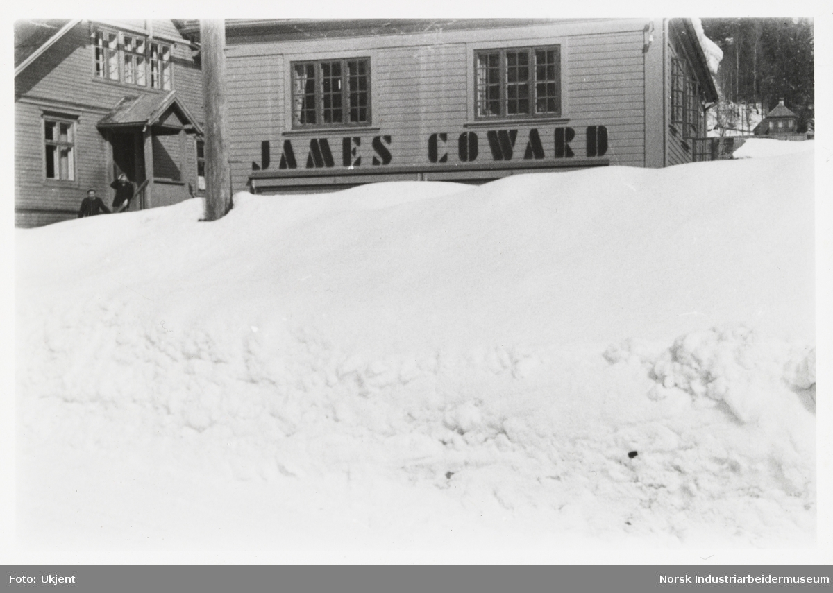 Mye snø i Sam Eydes gate foran butikkfasaden til jernvarehandelen James Coward. Til venstre sees to mennesker gå ned trapp