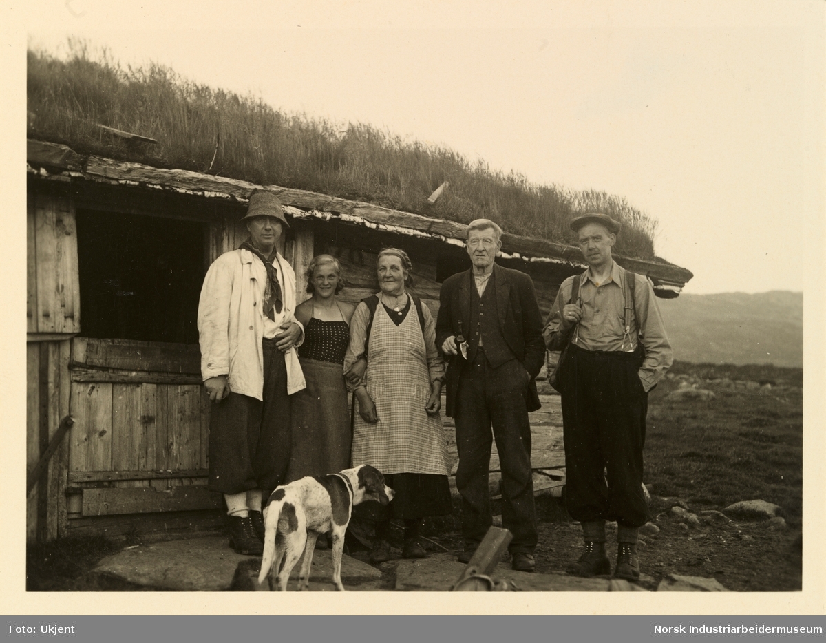 Tre menn, to damer og en hund foran hytte i fjellet