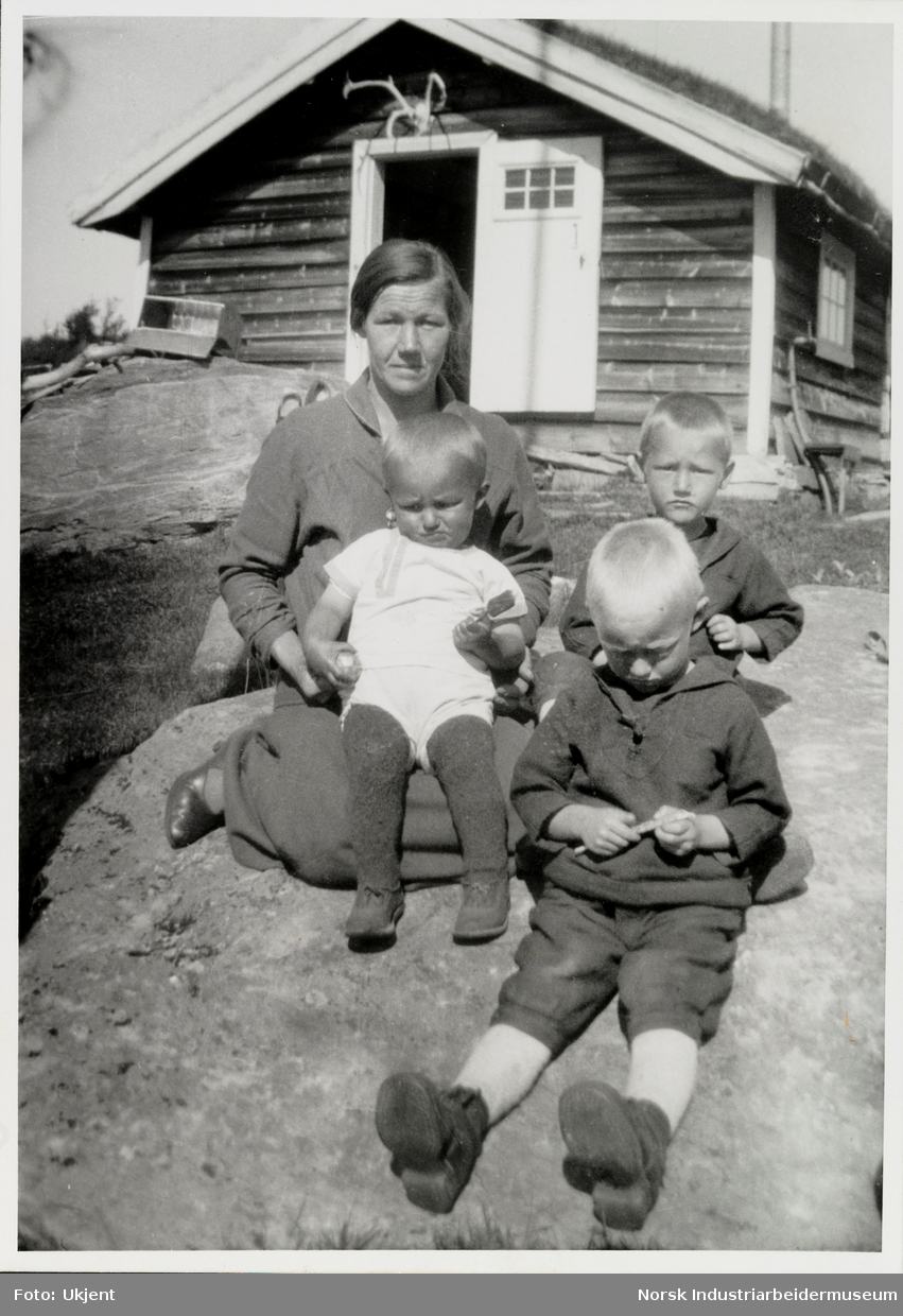 Kvinne med tre små guttebarn sitter på stein foran uthus med gevir over døråpning