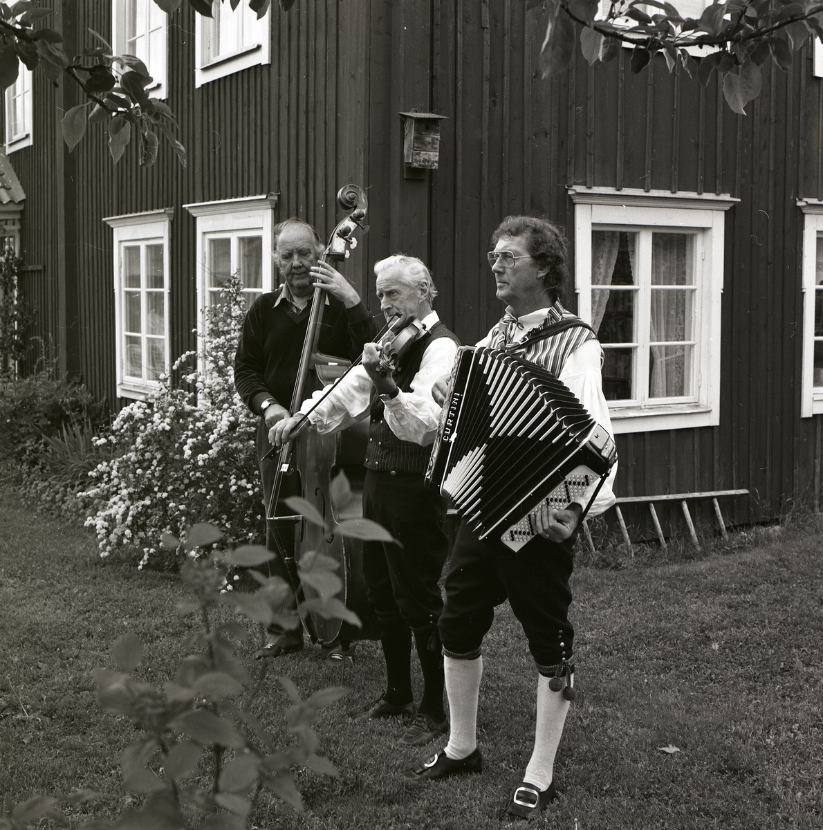 Tre män står på gräset intill ett hus och spelar på sina instrument, Norrgården den 21 juni 1986.