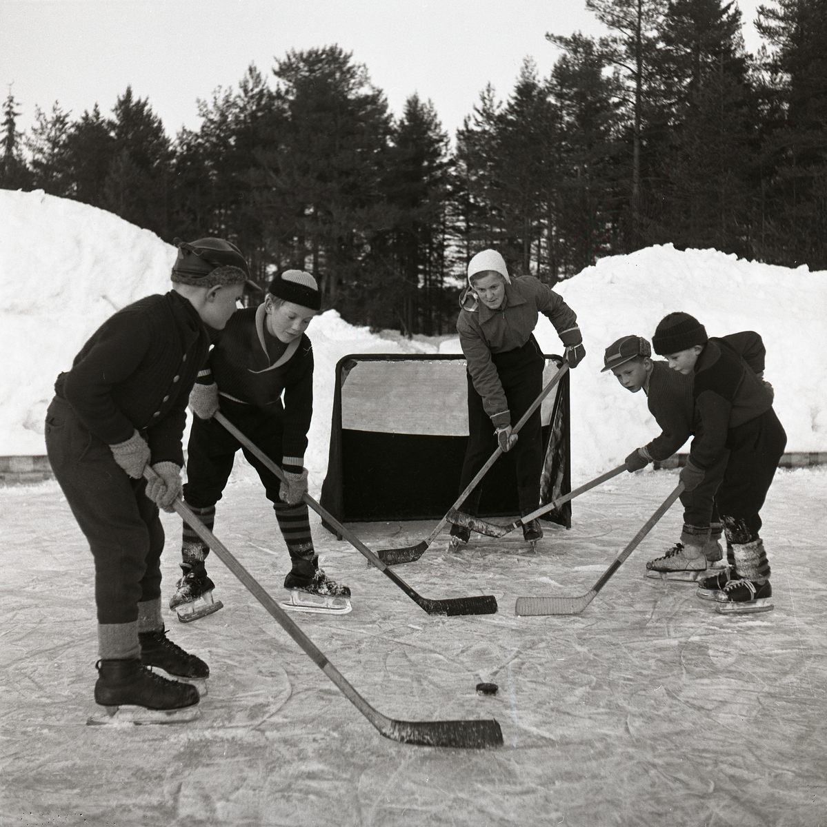 Det är några pojkar på isplanen och spelar ishockey, dom har skridskor, ishockeyklubbor, puck och en målbur. I bakgrunden är det snöhögar, skog och himmel. Ishockey Glösboplanen 27 januari 1959.