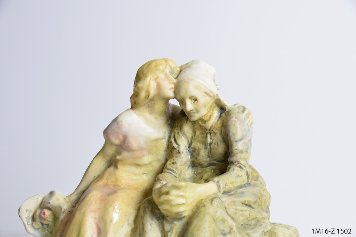 Skulptur som visar två kvinnor, en ung som håller om den äldre. Den unga håller även en bukett blommor i den högra handen.