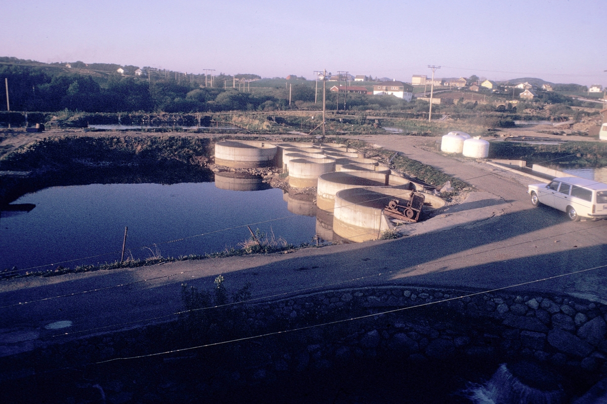 Oppdrett, Egeland, 1974 : I forgrunnen er flere jorddammer og en del betongkummer. Bak i bildet til høyre ser vi broen som krysser elva. Dambruket ligger like ved elva og får vannet sitt derfra.