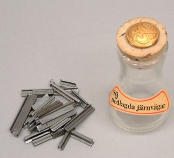 Liten glasflaska innehållande små bitar av skalaräls.
Gul etikett med texten "SJ nedlagda järnvägar". På korken är fastsatt en uniformsknapp från 1973 års reglemente.