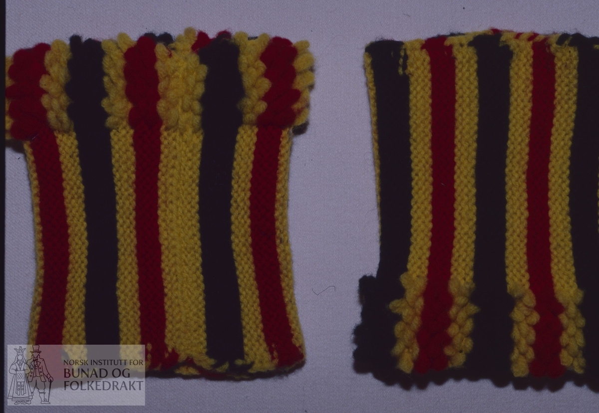 Rillestrikka rett i striper med gult, rødt og svart firetrådet ullgarn. Hver stripe har tre riller. I ene enden er det strikka inn løkker, 4 løkker som er strikka inn ved annenhver maske. Høgde ring:        ca 10,5 cm. Bredde løkkekant:  2,5-3,0 cm.