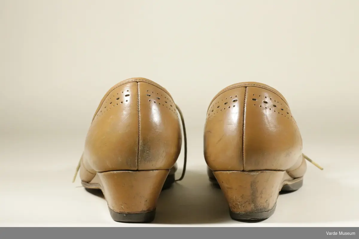 Brune damesko i skin. Skoen har en liten hel. Det er skolisser i brunt tøy, for å knytte skoen. Skoen er dekorert med små hull på midten av skoen, og langs kanten. Skoene er delvis slitt inni og på sålene. Skoen har skostørrelse 39. Skoene er av finsk fabrikat. 