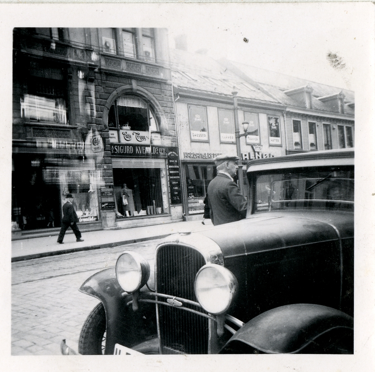 Fra familiealbum. Fotografi tatt fra gaten mot ulike forretninger, nummer 21 og 18 i Olav Tryggvasons gate Trondheim.
Bilen er en Oldsmobile 1932 modell.