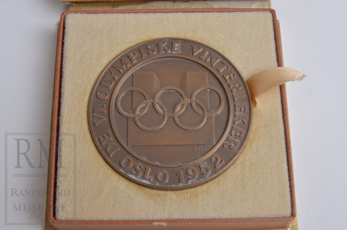 Medaljong i etui. De olympiske ringene i relieff foran Oslo rådhus. Tekst langs kanten. Snøkrystall i relieff med tekst på baksiden. Etui i brunt med olympiske ringer i gull. Gulhvit inni. Bånd til å heve medaljen med.