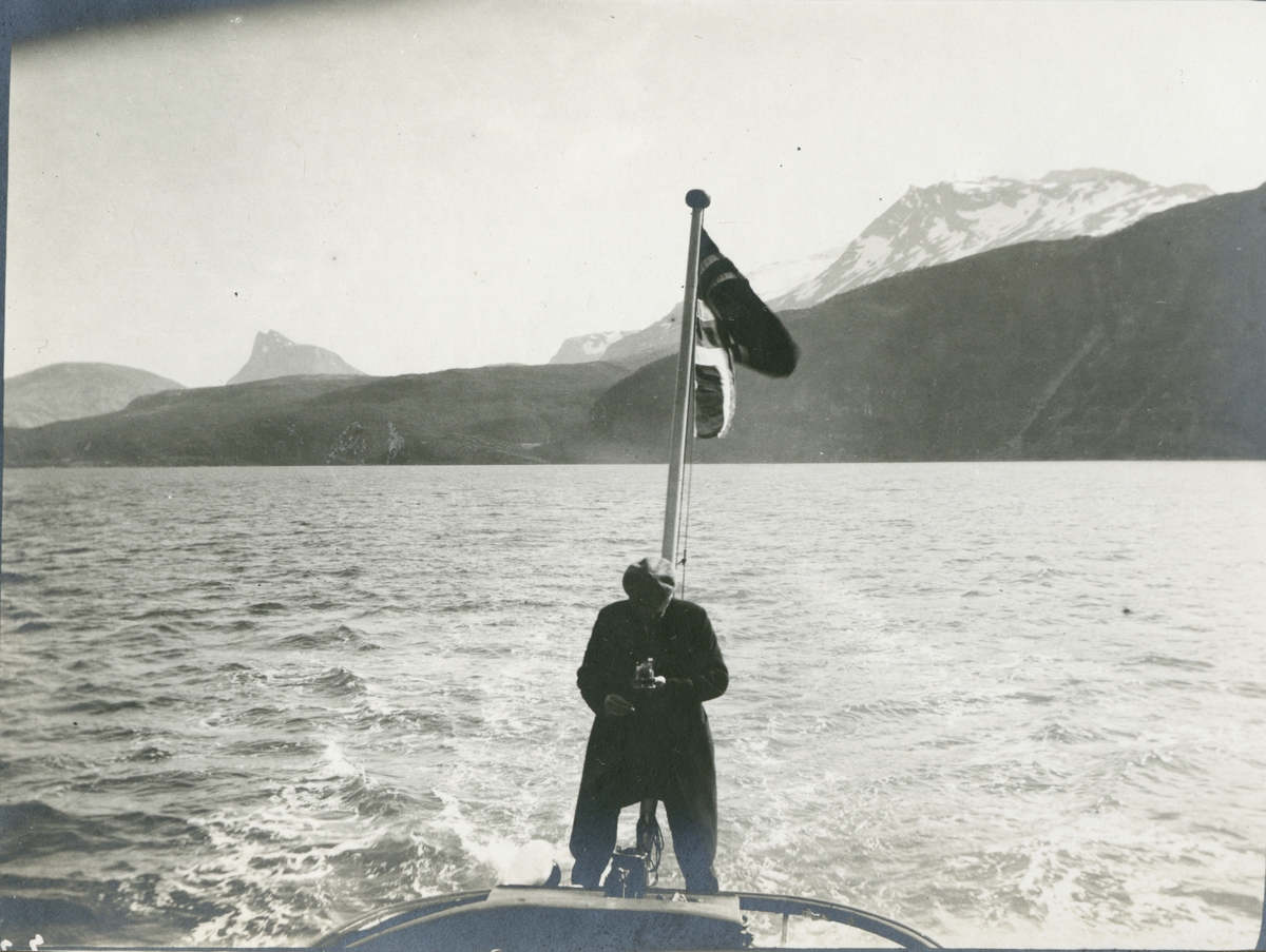 Bildtext: "Ploman fotograferar"
Ombord ett mindre norskt fartyg i en fjord. Man med bälgkamera står i aktern och fotograferar.