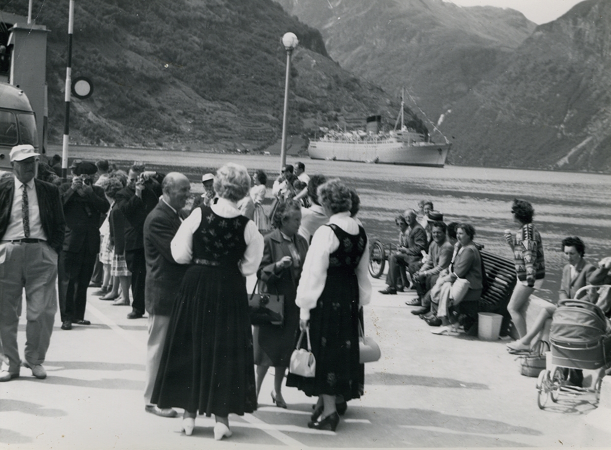 Motiv av en folkemengde på fergekaia i Geiranger. På fjorden ligger et større cruiseskip RMS "Caronia" fra selskapet Cunard Lines. I forgrunnen står to kvinner i bunad.
