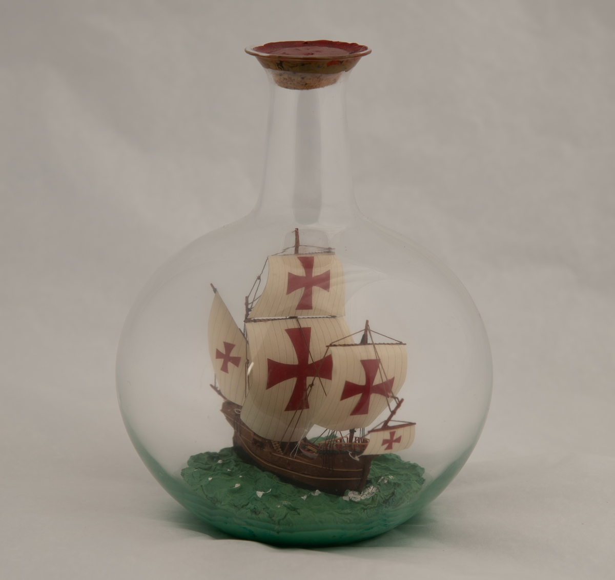 Flaskeskute i kuleformet flaske. Skuta er: "Santa Maria" , 1492 , en karakk , som var flaggskipet til Columbus.  Brunt skrog med lyse striper , seil med malteserkors i rødt.