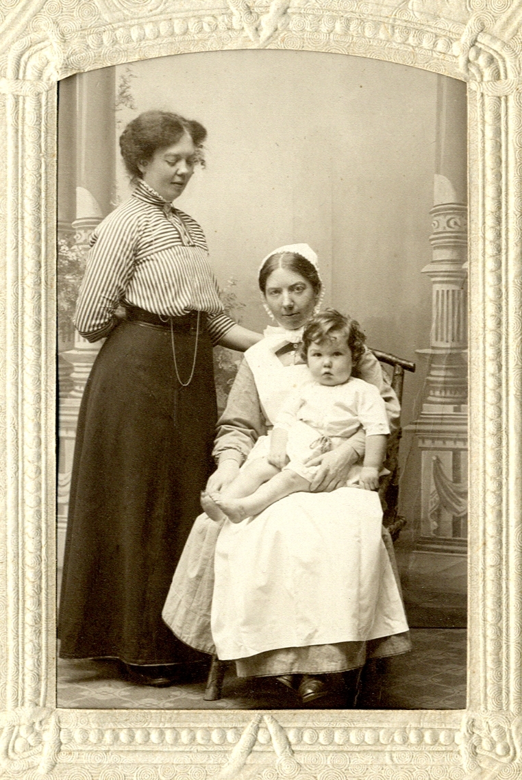Foto av två kvinnor och en liten flicka. Flickan sitter i knäet på den ena kvinnan, som är klädd i barnsköterskeuniform.
Helfigur. Ateljéfoto.