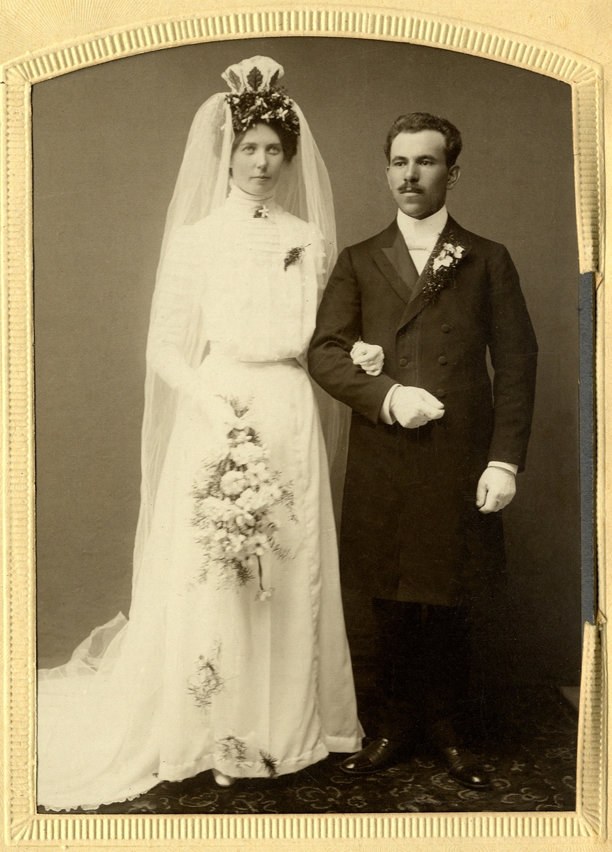 Foto av ett brudpar. Hon är klädd i vit brudklänning med slöja. Han är klädd i bonjour m.m.
Helfigur. Ateljéfoto.