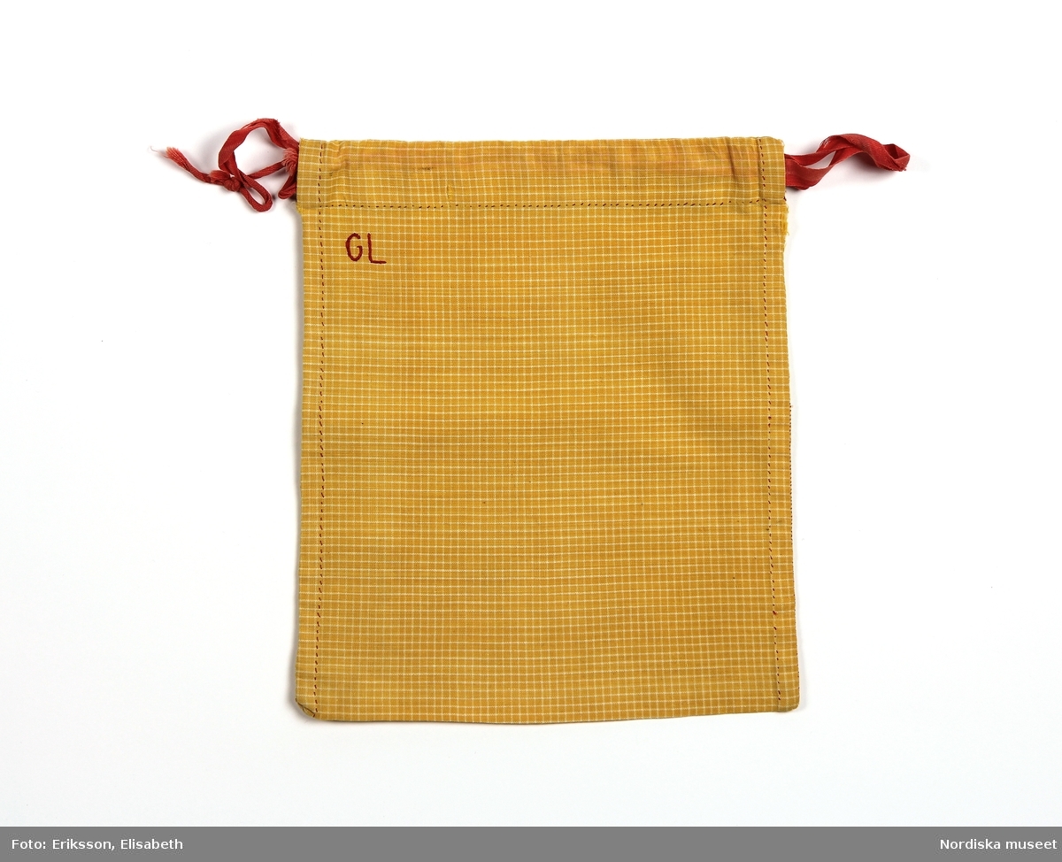 Påse sydd av gult tyg med tunna vita rutor. Handsydd med röd tråd. Dragsko och röda bomullsband. Monogram. 
/Fiffi Myrström, 2014-02-24