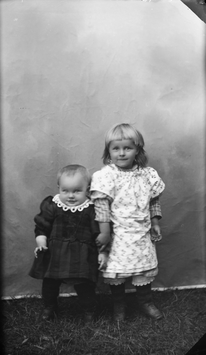 To søstre antatt ca 1 og 3 år. Den yngste i mørk kjole med blondekrage og den eldste med rutemøsntret sjorte og lys kjole med anker-motiv