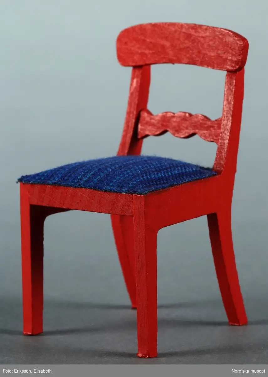 a) Ett köksbord och b-e) fyra stolar, alla av rödmålat trä, men av olika modell. b-c) Två av stolarna är raka och funktionella trästolar, troligen från 1950-talet, medan d-e) två har allmogekaraktär med utskuren dekor i ryggpartiet och blårandig klädsel. De senare är förmodligen från 1970-talet. Bordet är hemtillverkat av givarens far medan stolarna är tillverkade av Lundby. Hör till frukostrummet på första våningen i dockskåp NM.0331721+.