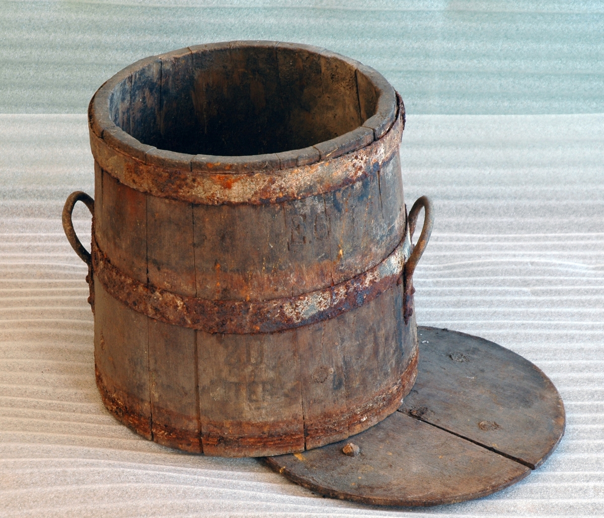 Sylinderformet, lagget bøtte av tre med band av jern. Bunn, mangler topp. Et håndtak av jern på hver side.