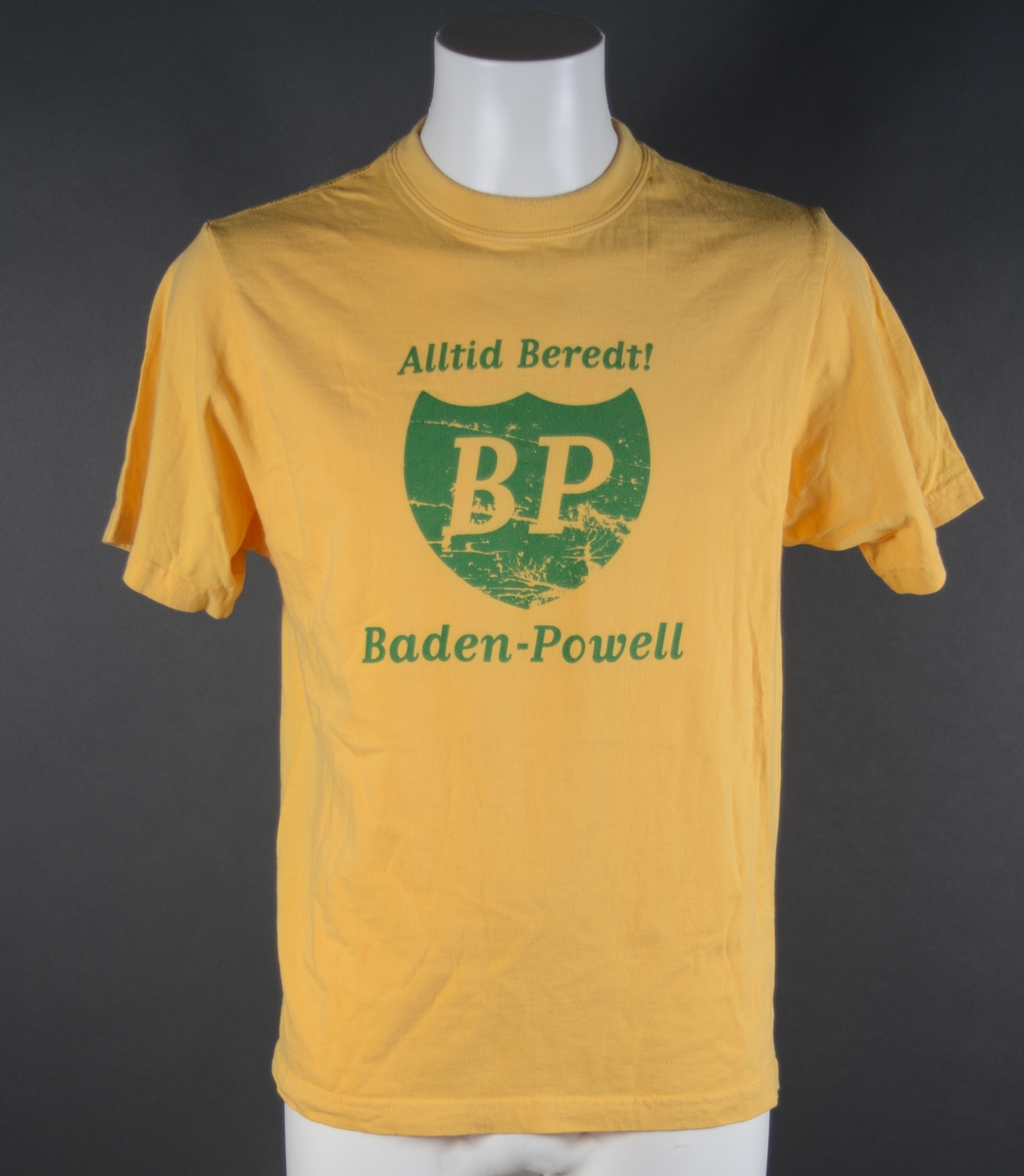 En T-skjorte med påtrykt tekst. Genseren er sydd av gult bomullsstoff som er elastisk. Den er i størrelse medium. Ermene er isydd. Halskanten er rund. Genseren har en søm i hver side, og nederst er det sydd opp en kant. I nakken er det sydd på en merkelapp, se "Påført tekst/merker". I front er det påtrykt dekor. I midten er logoen til BP (British Petroleum). Over står det "Vær beredt" som på engelsk er "Be Prepared" = BP. Under står det Baden-Powell som også passer i ordspillet BP. Dekoren er i grønt.