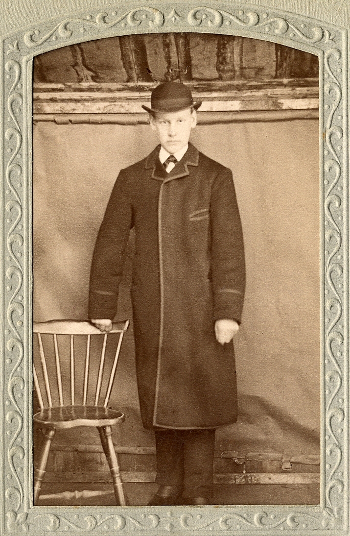En okänd ung man i rock och hatt (plommonstop/kubb). Han håller ena handen på ryggstödet till en pinnstol. På baksidan av fotot finns antecknat: "13/2 1886". 
Helfigur, en face. Ateljéfoto.