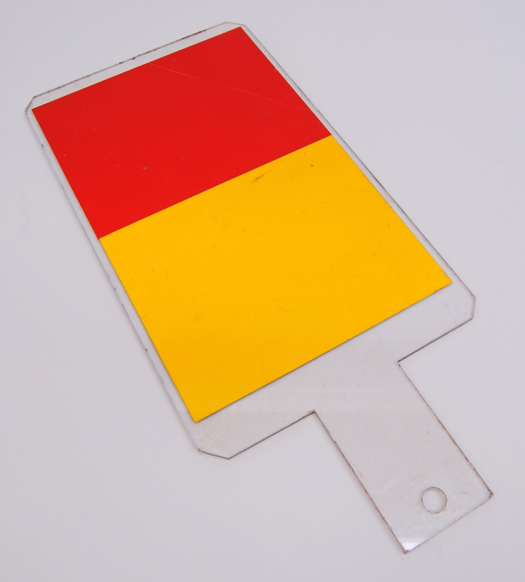 Slutsignalsskärm av genomskinligt plast. Den är formad som en stående rektangel med beskurna hörn. I ena änden finns ett handtag med ett hål i ena änden. 

Den ena halvan av skärmen är gul och den andra röd.
