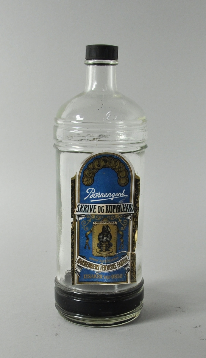 Flaske av glass med svart plastkork for skriveblekk. Etikett av papir med bilde av en bjørn i sentrum, med tekst og dekor i hvitt og gull på blå bakgrunn.