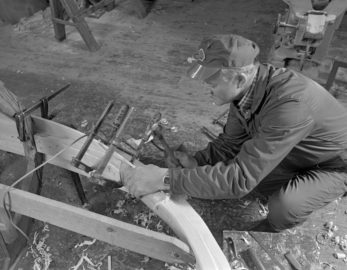Bygging av fløterbåt i Glomma fellesfløtingsforenings verksted Breidablikk ved kommunesenteret Flisa i Åsnes i november 1984. Båttypen ble for øvrig kalt «Flisabåt», men denne modellen var egentlig utviklet av John Dybendal (1904-1985) omkring 1930. Målet var å kunne levere en robåt som passet i elva Flisa. Dybendal bygde sjøl slike båter fram til 1949. Glomma fellesfløtingsforening hadde vært en viktig kunde hos Dybendal, og da han gav seg som båtbygger ble virksomheten flyttet til denne virksomhetens verksted for Solør-regionen, hvor båtbygging ble vinteraktivitet for et par av karene som hadde vassdraget som arbeidsplass sommerstid. Det var maler til alle båtdeler, og skroget ble bygd på tverrmaler som ble festet til kjølen. Dette var hjelpemidler som gjorde det mulig for fløtere med reletivt liten håndverkserfaring å bygge denne båttrypen og oppnå den standardiserte formen som fløterkameratene var vant med. De fleste flisabåtene fikk jernspanter, for det var krevende å finne de krokvokste emnene som gav de beste spantene. Da Norsk Skogbruksmuseum dokumenterte båtbygginga ville karene likevel bruke trespanter. Da dette fotografiet ble tatt spikret Lars Bernhard Olastuen (1927-1998) den første bordgangen til kjølen, etter først å ha festet den til de nevnte spantmalene ved hjelp av tvinger. Flisabåtene ble, som dette bildet viser, bygd løst. Kjølen var ikke fastspent i verkstedet, og den utferdige båten kunne dermed vippes fra side til side slik det passet under ulike faser i arbeidet.

Båten som var under bygging da dette fotografiet ble tatt ble seinere overlatt til Norsk Skogbruksmuseum, der den fikk nummer SJF.07823 i gjenstandssamlinga. Den er 518 centimeter lang og 153 centimeter bred. Odd Jensen har seinere lagd oppmålingstegninger, som også finnes i museets arkiv.