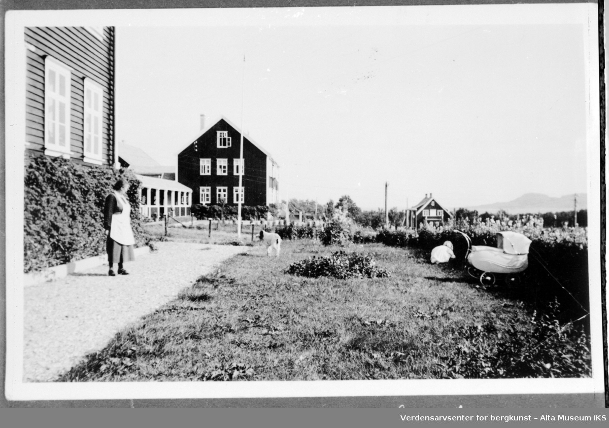 Doktorgården i Talvik med Tuberkolosehjemmet i bakgrunn. Bildet viser en kvinne og et barn som leker i en busk, en hund og en barnevogn.