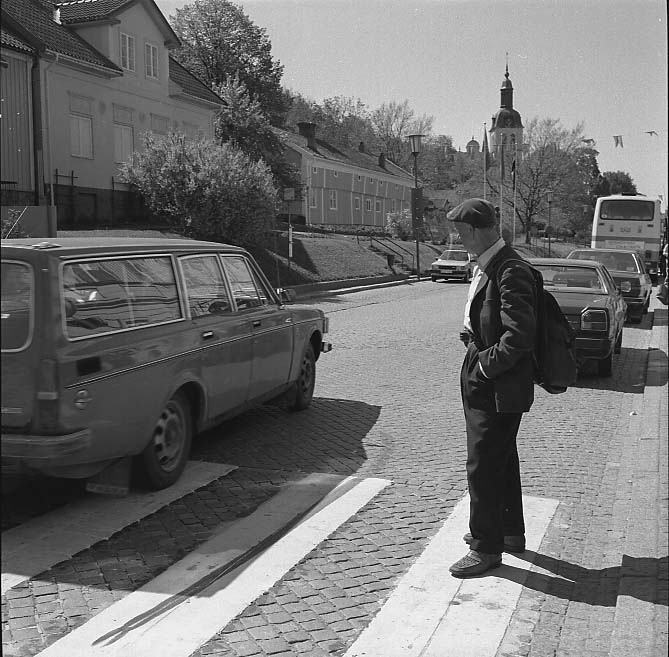 En man i keps står ett steg ut i gatan på ett övergångsställe. En Volvo kombi passerar precis och mannen tittar efter den. På andra sidan gatan syns kvarteren Hjorten. Mannen på bilden är Erik Karlsson, mer känd som "Erik i Hägnen".