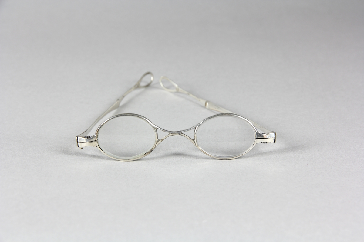 Glasögon, mindre glas och smala bågar av silver. Två ledade skalmar som utåt avslutas med droppformade öglor.