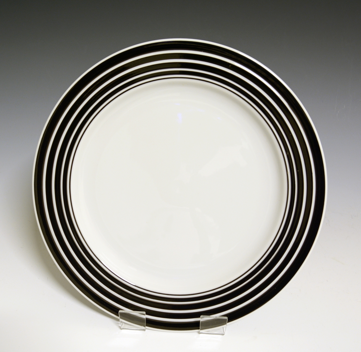 Middagstallerken av porselen med hvit glasur. Fanen dekorert med med sorte striper som øker i bredde mot fanens ytterkant. I fanens gods riller. 
Modell: Saturn