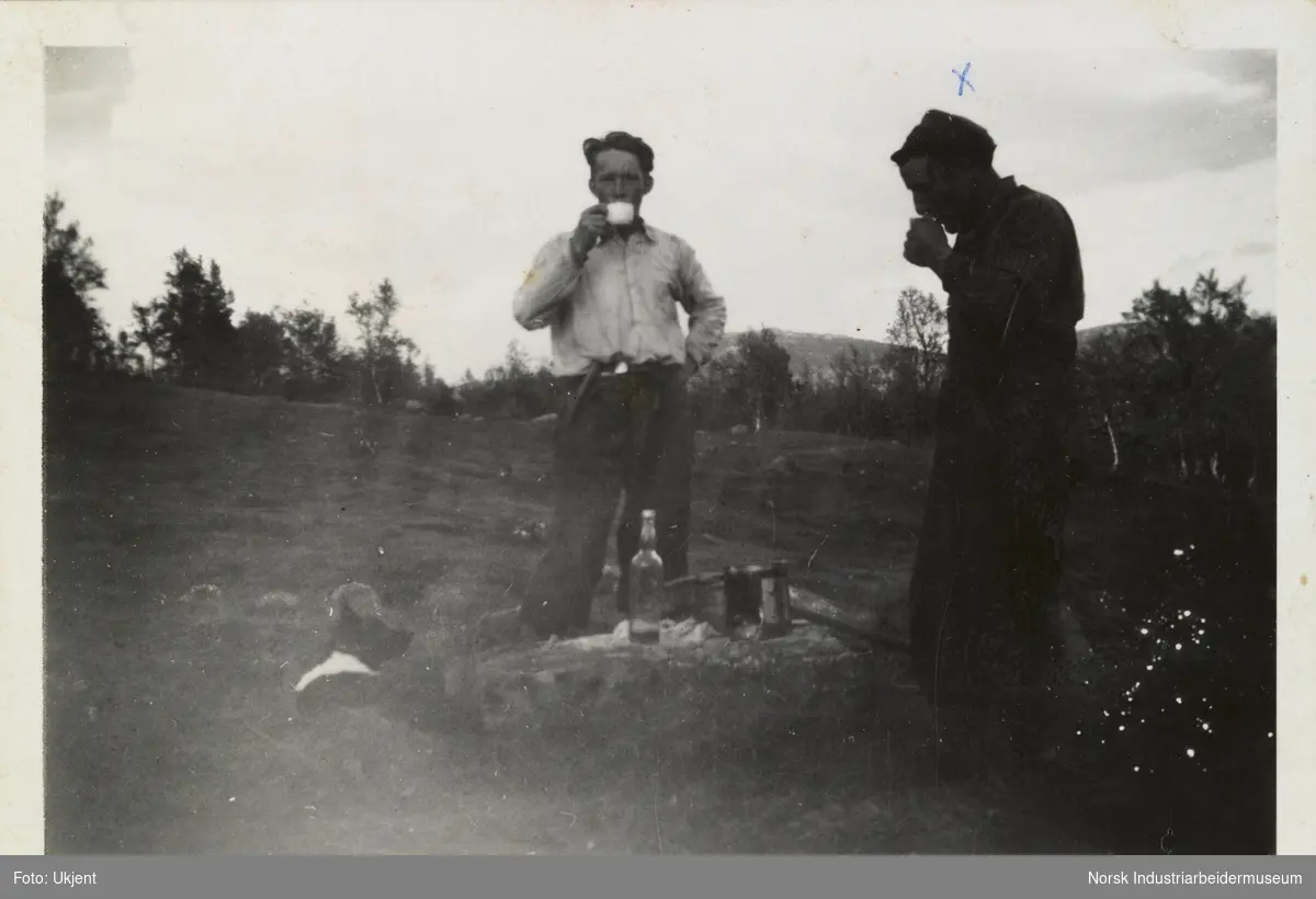 Tarjei Skinnarland og en mann står på beite og drikker fra kopp. På steinen mellom dem står flaske og en kjele