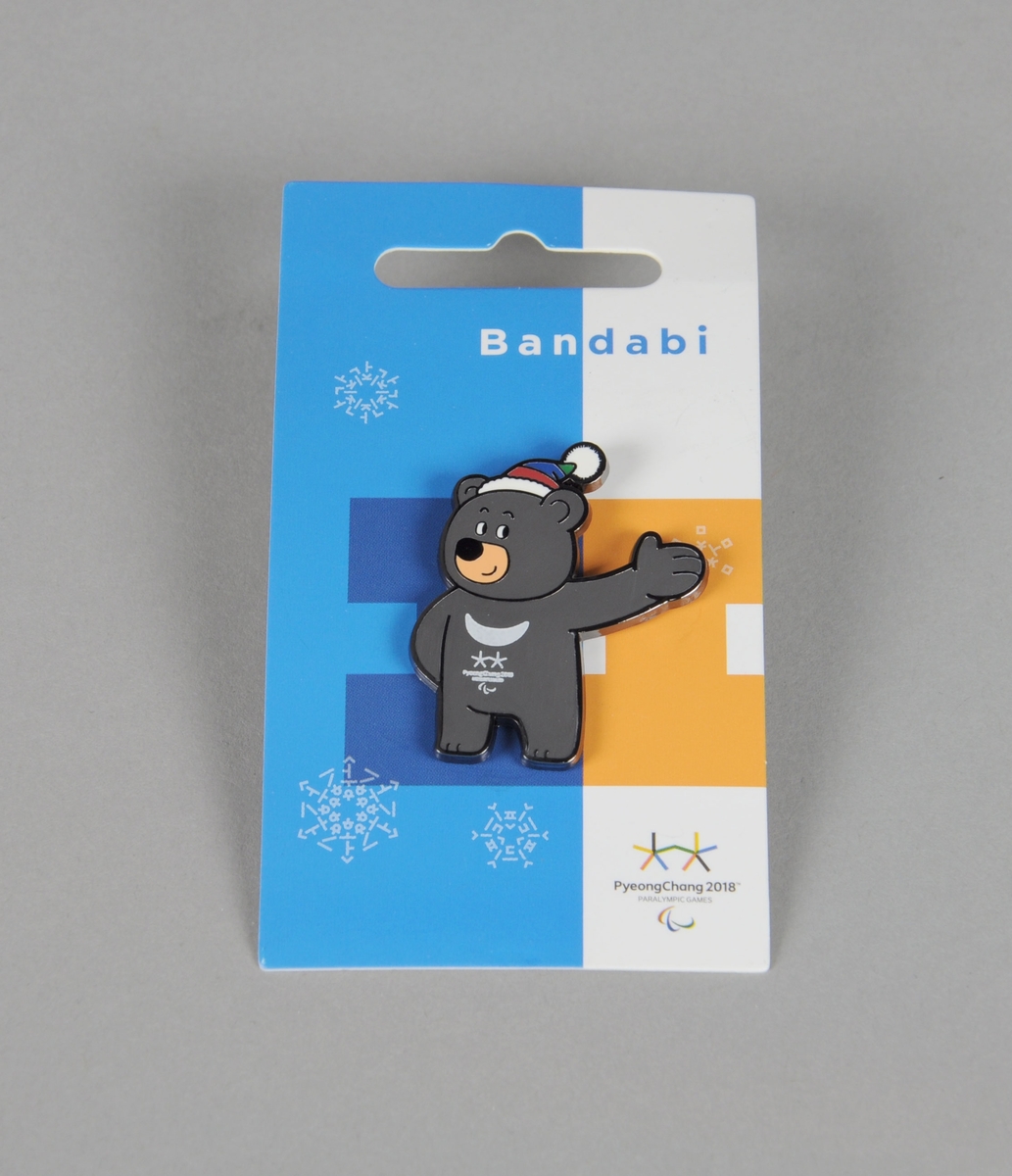 Jakkemerke formet som maskoten Bandabi med emblemet for vinter-Paralympics i PyeongChang 2018. Merket ligger i orgiginal emballasje merket navnet på maskoten og embelet for Paralympics. På baksiden av emballasjen er det et hologram med emblemet for lekene med et unikt nummer