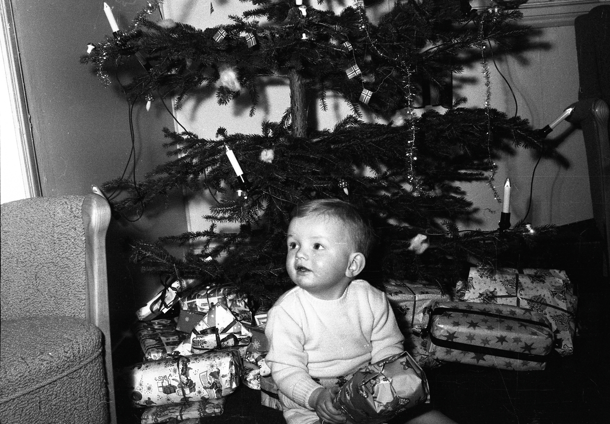 Gutt med julegaver foran juletre.