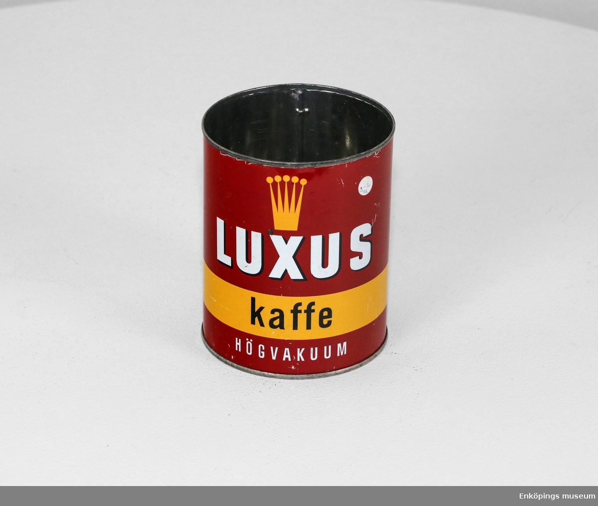 Röd plåtburk med gul bård. Burken är försedd med en krona följt av texten: "Luxus kaffe högvakuum". På burkens sida kan man läsa om de tre faktorerna som påverkar smaken.