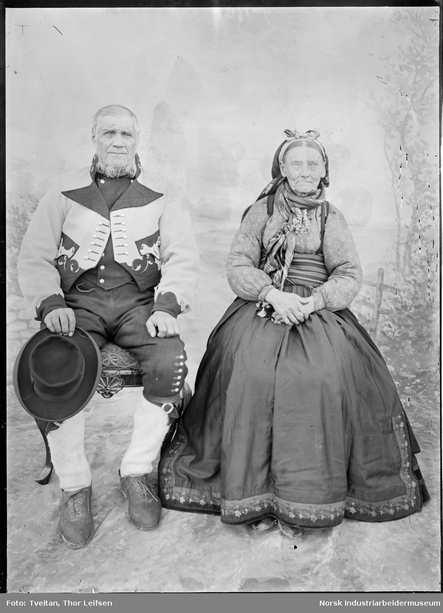 Ateliefoto. Eldre ektepar sittende på stol i øst-Telemark bunad. Damen i beltestakk med hårbånd og skaut. Mannen i gråtrøyebunad med knebukse.