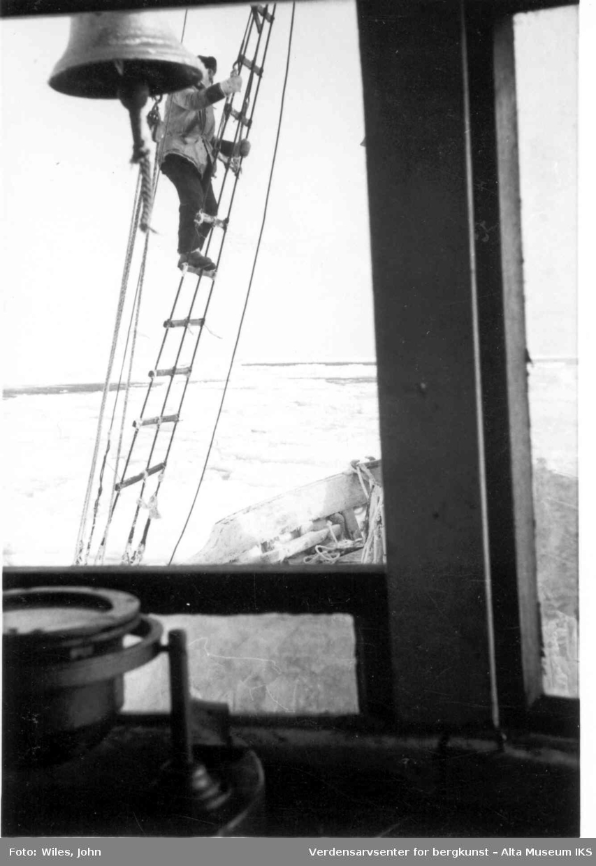 Mann klatrer i taustigen opp masten på sshavskuta Polis. Bildet er tatt fra innsiden av styrhuset. Mannens ansikt er skult av en bjelle. 
I bakgrunn strekker isen seg utover havet.
