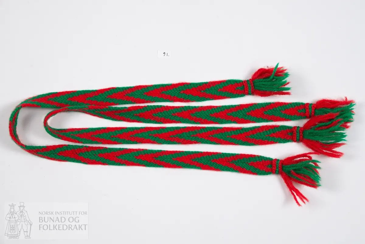 Sokkebånd.
Et par fingrede sokkebånd av rødt og grønt ullgarn med dusk i hver ende.
