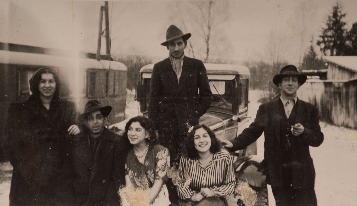 En grupp romska män och kvinnor är uppställda framför en bil. I bakgrunden syns en bostadsvagn.