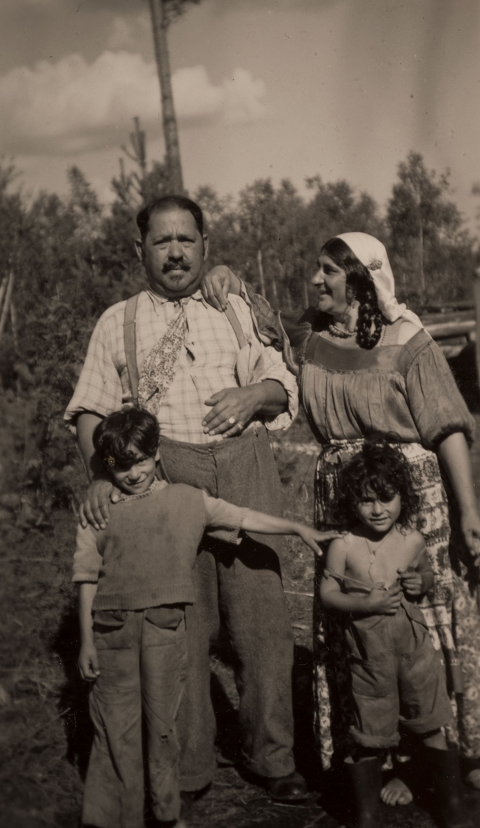 En grupp romer samlade för fotografering, en sommardag i Hofors 1950. Två barn står framför en kvinna och en man. Kvinnan har flätat hår och en "dikhlo", en huvudduk som markerar att hon är gift. På dikhlon sitter ett spänne.
