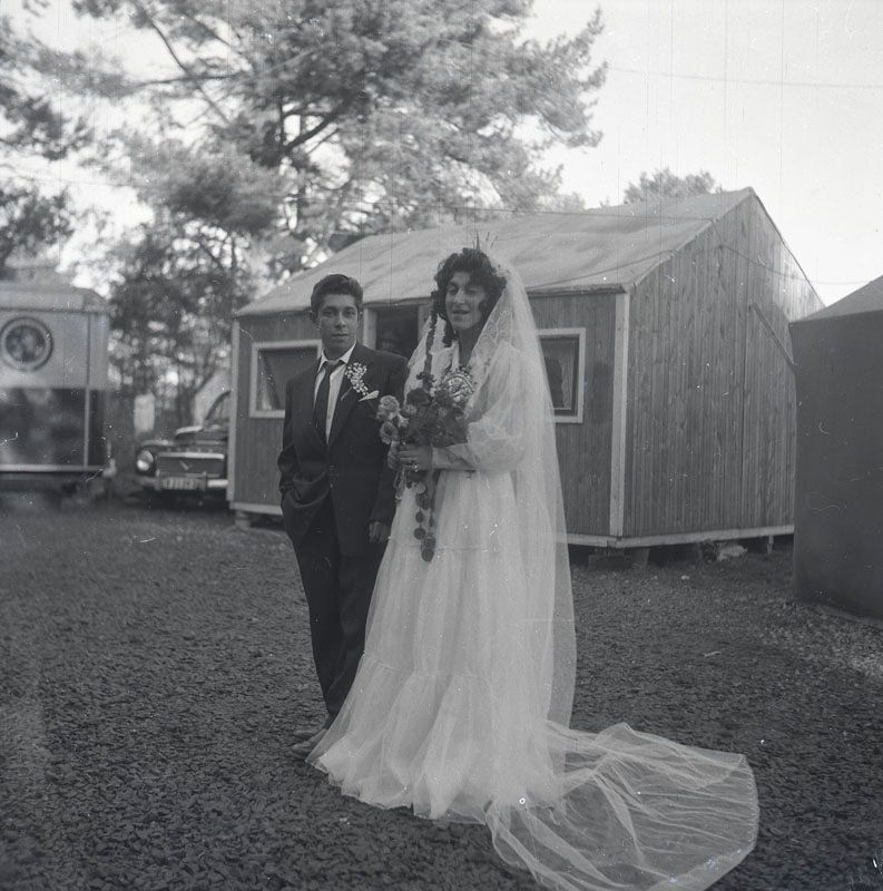 Nygift romskt par uppställda för fotografering vid bröllop i november 1958 i Nyköping. I bakgrunden syns uppställda vagnar, baracker och bilar i lägret.