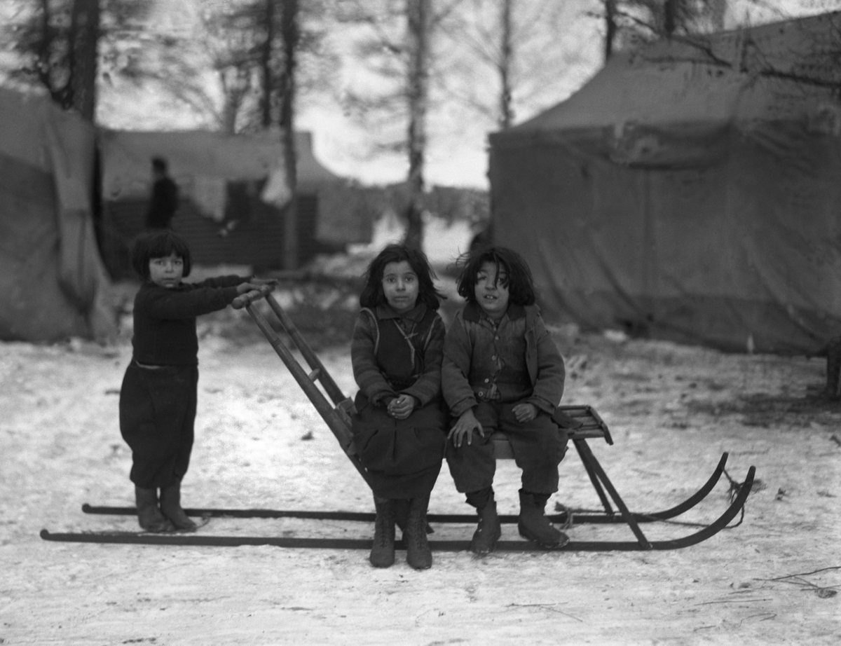 Tre romska barn sitter på en sparkstötting. I bakgrunden syns tälten i lägret som barnen bor i. Landskapet runt om dem är täckt av snö.