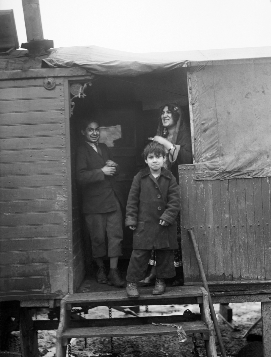 En romsk kvinna och två barn står i dörröppningen till en bostadsvagn i ett läger i Gröndal. Kvinnan bär en dikhlo.