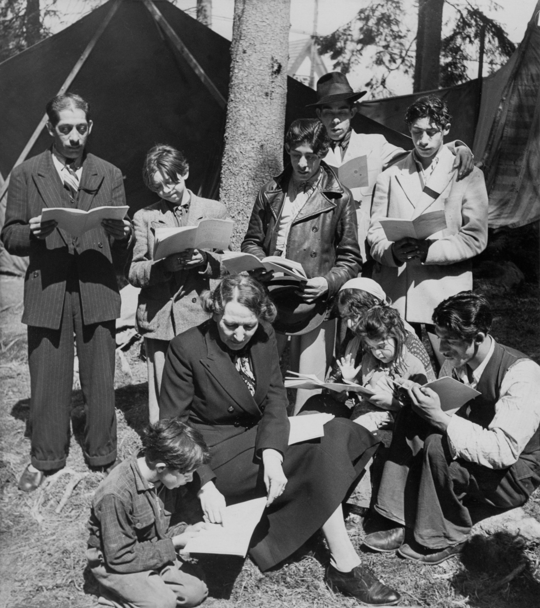 Här syns Sveriges första ambulerande skola för romer, år 1946. I mitten av bilden sitter klassens lärare omgärdad av elever i alla åldrar som läser i studiematerial. Bakom dem syns tältdukar.
