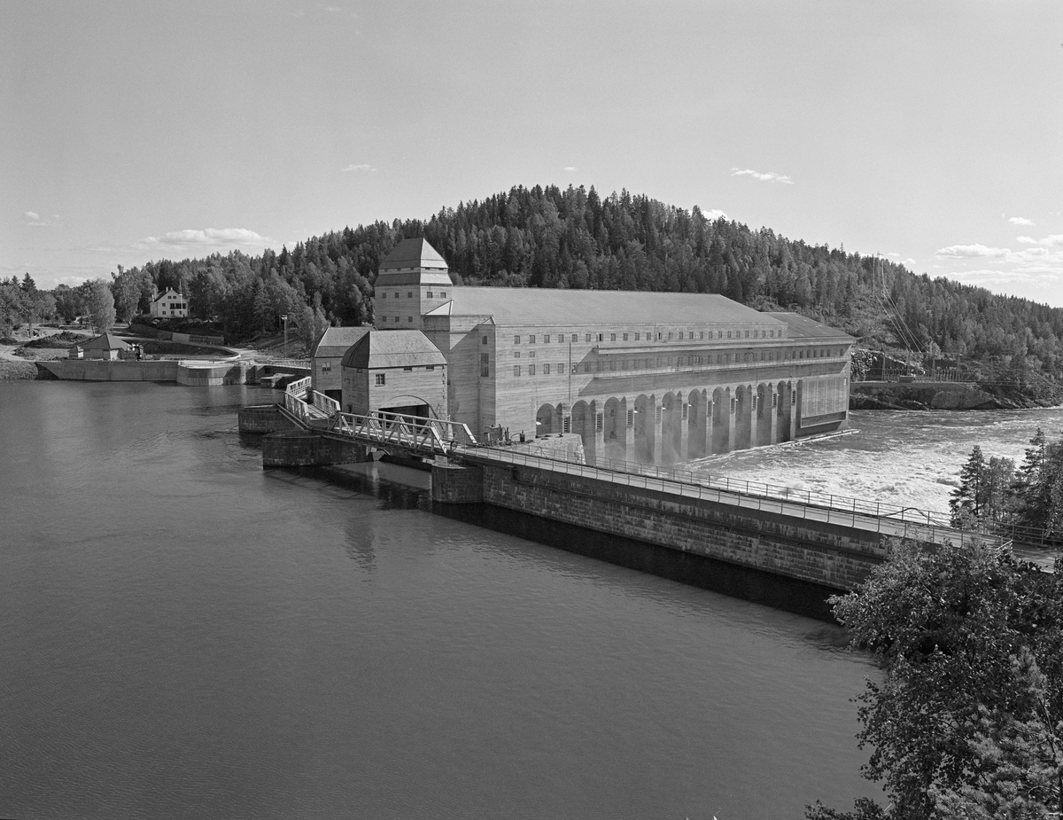 Mørkfoss/Solbergfoss kraftstasjon i Askim, sett fra nord, med kraftverksdammen i forgrunnen, kraftstasjonen i mellomgrunnen og Tyrihjellåsen i bakgrunnen. Dette kraftverket ble bygd i perioden 1917-1924 med staten (som eide ovenforliggende Mørkfoss med 10,5 meters fall) og Oslo kommune (som eide mellomliggende Vittenbergfoss, Skråperudfoss og Halfredsfoss, som hadde 5 meters fall ved lavvann). Dammen ble plassert på terskelen i den opprinnelig todelte Solbergfossen, som inntil da hadde hatt cirka 2,5 meters fallhøyde ved lavvann.  Etter at det ble reist en massiv betongdam ved Solbergfossen kunne man heve vannspeilet cirka 25 meter, på nivå med den ovenforliggende innsjøen Øyeren, som dermed ble en del av reservoaret til dette kraftverket. For å kunne gjøre det store inngrepet i vassdraget som dette kraftverksprosjektet innebar, måtte utbyggerne forhandle med Fredrikstad Tømmerdirektion, som hadde hevd på å bruke Nedre Glomma som transportåre for tømmer. På dette tidspunktet var det vanligvis 6-7 millioner stokker årlig som passerte fossen på veg mot sagbruk og papirfabrikker i området ved Fredrikstad og Sarpsborg. Forhandlingene om hvordan dette skulle løses involverte tung ingeniørfaglig og juridisk ekspertise. Det gikk halvannet tiår før man endte med en tunnelløsning som begge parter var fornøyde med. Kraftstasjonen ble tegnet av den kjente arkitekten Bredo Greve (1871-1931). Da kraftstasjonen ble satt i drift (1924) hadde den 7 Francis-turbiner. I løpet av 1930-åra ble det installert ytterligere tre. Det vi ser i forgrunnen på dette fotografiet er dammen, som hadde tre overløpsåpninger med fagverksbruer over. Inntaksbassenget befant seg i det området som på dette bildet ligger bak kraftverksbygningen. Fløtingstømmeret ble ført forbi kraftverket via ei renne på nordvestsida av vassdraget med inntak omtrent der hvor fotografen sto da dette fotografiet ble tatt. Fløtinga ble avviklet etter 1985-sesongen. Produksjonskapasiteten ved anlegget er seinere betydelig utvidet - jfr. fanen «Opplysninger».