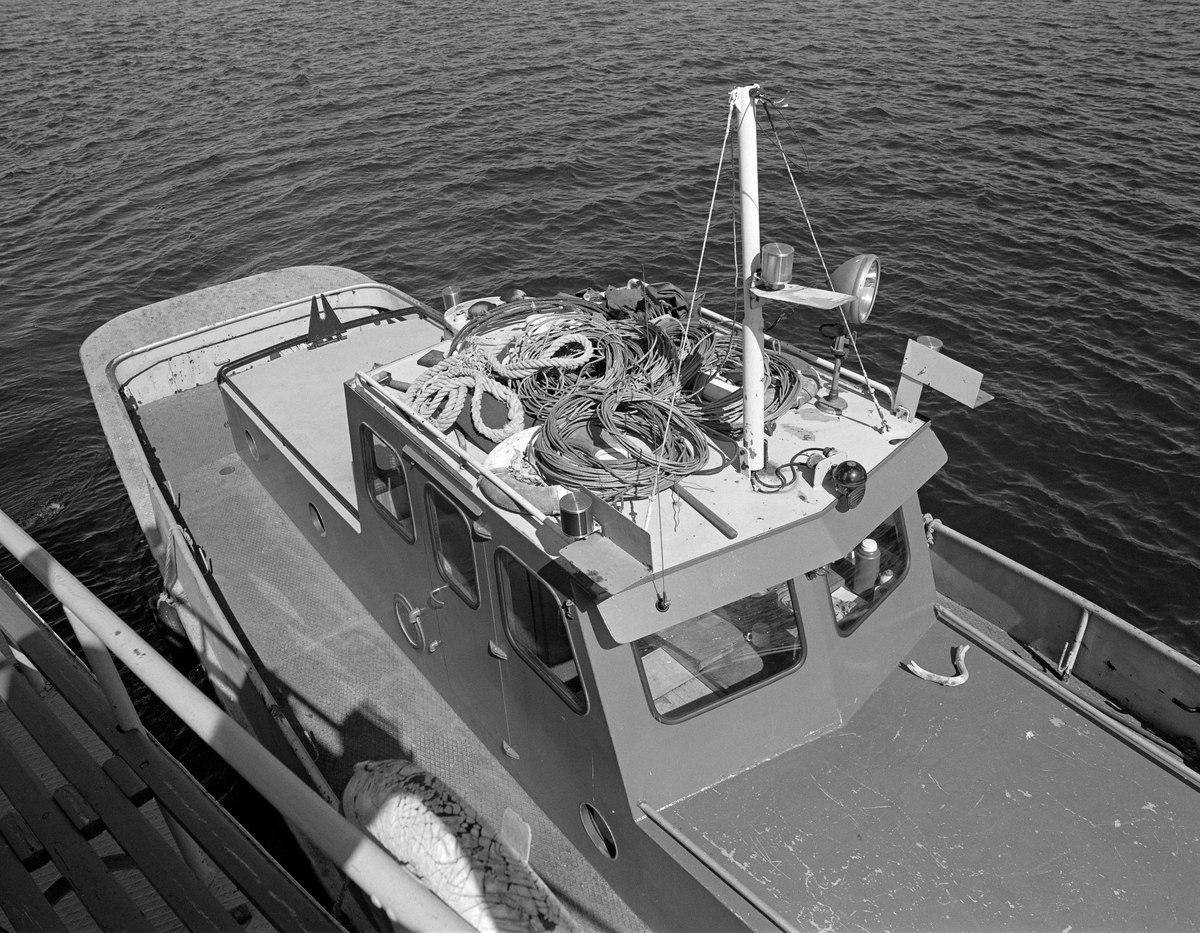 Styrhuset på den lille slepebåten Rauma, som var hjelpebåt under slepinga av tømmer i den nordre delen av innsjøen Øyeren.  Her var det et deltaområde hvor slepene måtte følge en trang og svingete djupål.  Den kraftige slepebåten Mørkfos (jfr. SJF-F. 005195) ble brukt som trekkraft, mens den mindre båten Rauma (se også SJF-F. 005198) gikk bak for å sikre at bakenden av slepet fulgte den oppmerkete seilingsleden.  Dersom deler av slepet gikk på grunn eller andre uhell inntraff, oppsto det behov for å binde samle tømmeret igjen og binde i hop slepet på nytt.  Det er bindsel til slike reparasjoner som ligger på taket av Raumas styrhus.  Det ble særlig brukt «grimer» av den typen som ble brukt på «soppene» (tømmerbuntene) fra lenseanlegget på Fetsund.  Fotografiet er tatt fra dekket på den større slepebåten Mørkfos. 