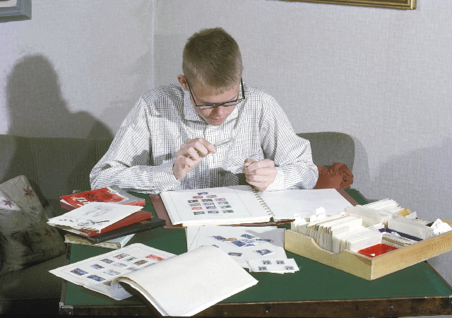 Seriebild M 14. Ung frimärkssamlare studerar frimärken med
förstoringsglas. Han är omgiven av frimärksalbum och kataloger.